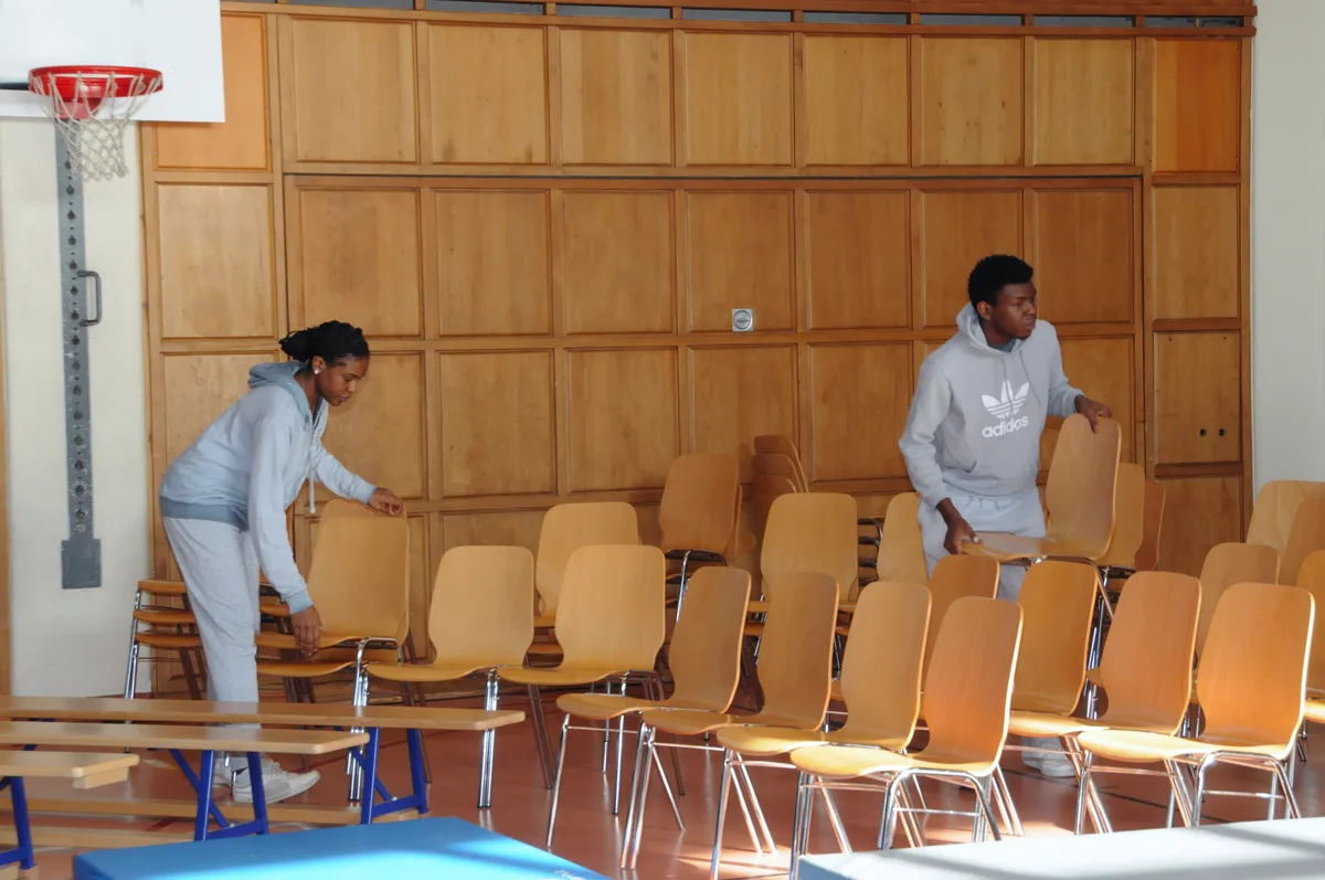 Man sieht eine Frau und einen Mann beim Aufräumen von Stühlen in einer Turnhalle.
